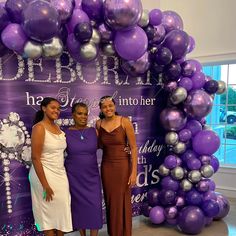 three women standing in front of a purple and silver balloon arch with the words happy birthday on it