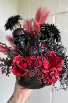 a hand holding a black vase with red flowers and skulls on the wall behind it