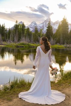 a woman in a wedding dress standing by a lake with mountains in the background at sunset