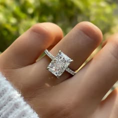 a woman's hand with a diamond ring on top of her finger, in front of some bushes