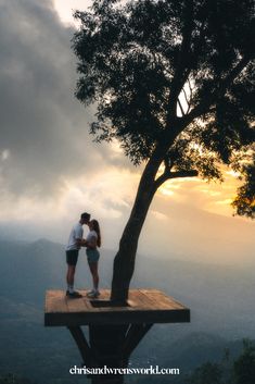 two people standing on top of a wooden platform next to a tree and mountains in the background