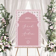 a pink and white wedding sign in front of floral arrangements with greenery on either side