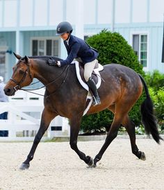 a woman riding on the back of a brown horse across a sandy field in front of a blue building