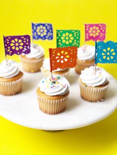 cupcakes with white frosting and colorful paper flags on a plate against a yellow background