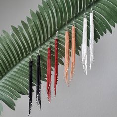 four pairs of earrings hanging from a green leaf with white beads and red, black, and white tassels