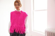 a woman standing in front of a window wearing a pink knitted sweater and black pants