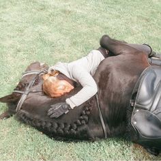 a woman laying on top of a black horse in the middle of some green grass