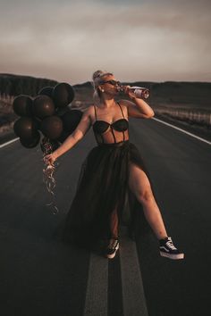 a woman in black dress sitting on the side of road holding balloons and drinking water