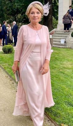 an older woman in a pink dress and hat standing on the grass near some stairs