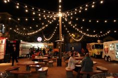 people are sitting at picnic tables in an open area with food trucks and lights strung overhead