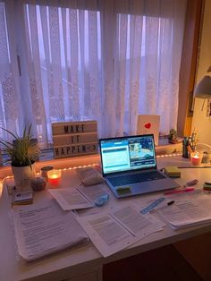 an open laptop computer sitting on top of a desk next to papers and candles in front of a window