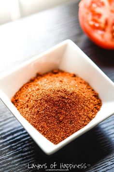 a small white bowl filled with red seasoning next to a tomato on a wooden table