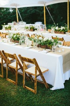 a long table set up with white linens and wooden chairs for an outdoor wedding reception