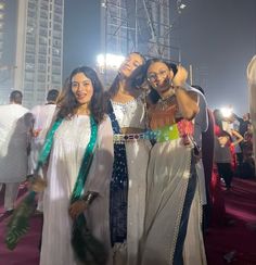 three women standing next to each other in front of tall buildings at night with lights on