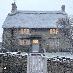 a stone house covered in snow next to a fence and gate with lights shining through the windows