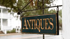 an antique sign hanging from the side of a white building with trees in the background