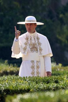 a man in a white outfit and hat making the peace sign with his hand while standing on top of some bushes