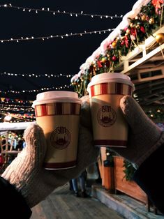 two cups of coffee being held up by someone's hands in front of christmas lights