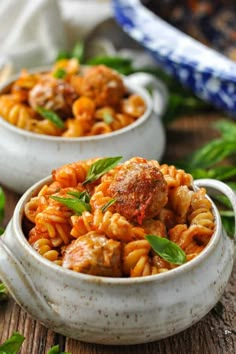 two bowls filled with pasta and meatballs on top of a wooden table next to green leaves