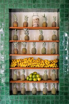 pineapples, limes and bananas are arranged on shelves in a green tiled room