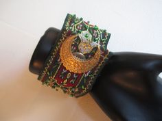 This gorgeous cuff measures 2 1/8 inches wide at the widest point and 7 1/4 inches long closed. The base brocade ribbon is beautiful with hints of gold lame, sprinkled with green glass beads. The center gold tone vintage piece is delicately carved and a vintage brass stamped crescent conceals the easy snap closure. Lined with emerald satin ribbon and gold lame. Unique and one of a kind! Handmade Green Bracelet For Celebration, Handmade Green Celebration Bracelet, Gold Cuff Bracelet For Festivals, Gold Embroidered Bracelets For Wedding, Traditional Green Adjustable Cuff Bracelet, Gold Embroidered Bangle Bracelet, Luxury Green Patina Cuff Bracelet, Luxury Vintage Green Cuff Bracelet, Vintage Adjustable Green Bangle