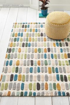 a white rug with multicolored squares on it next to a yellow pillow and potted plant