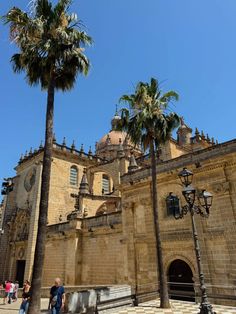 two palm trees in front of an old building with people walking around the courtyard and on the sidewalk