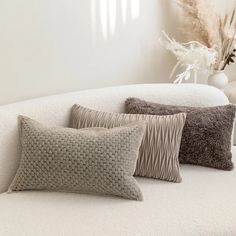 three decorative pillows on a white couch in a living room with a vase and dried pamodia