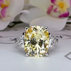 a fancy yellow diamond ring with three diamonds on it's sides and two white flowers in the background