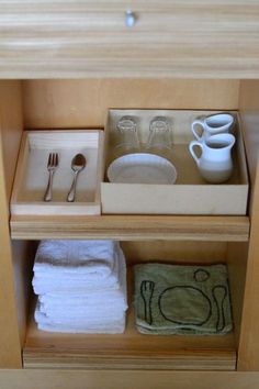 an open drawer with dishes and cups in it, next to folded towels on the bottom shelf