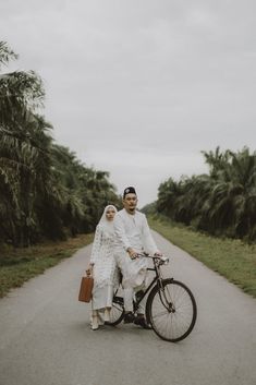 a man and woman riding on the back of a bike down a road next to palm trees