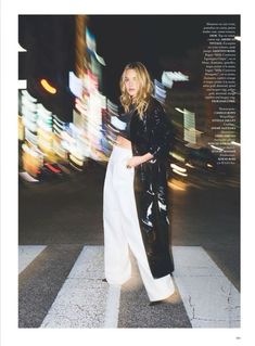 a woman in black jacket and white pants standing on the crosswalk at night with city lights behind her