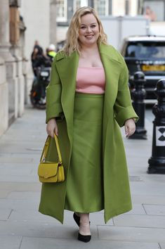 a woman in a green coat and pink top is walking down the street with a yellow handbag