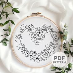 an embroidered heart with flowers and leaves on white fabric surrounded by greenery in the shape of a wreath