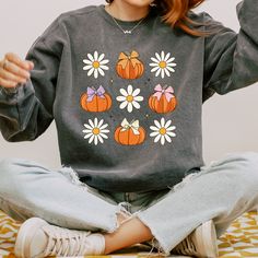 a woman sitting on the floor with her hands in the air while wearing a sweatshirt that has pumpkins and daisies on it