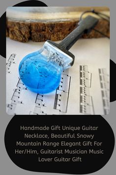 a blue object sitting on top of sheet music with words below it that read handmade gift unique guitar necklace, beautiful snowy mountain range elegant gift for her / him