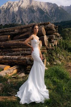 a woman in a white wedding dress standing next to some logs and grass with mountains in the background