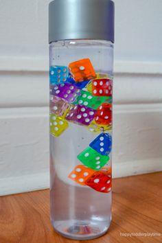 a water bottle filled with colorful dices on top of a wooden table next to a white wall