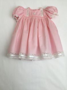 This size 3 mo. dress is made of pink, poly/cotton fabric.  Lace insertions and edgings are on the bodice, sleeves and dress bottom.  The dress is lined with white polyester cooton.  A white heart button is added to the bodice and the back is also fastened with 2 heart buttons.  The body of the dress has several rows of smocking, embedded with white beads.   The bottom of the dress has rows of ribbon and lace.  French seams are included for durability and an heirloom touch. The dress is lined. F Fitted Pink Cotton Dress, Cotton Short Sleeve Dress For Baptism, Pink Doll Collar Dress For Dress-up, Pink Dress With Doll Collar For Dress-up, Pink Smock Cotton Dress, Pink Cotton Smock Dress, Short Sleeve Dresses With Buttons, Cotton Baptism Dress With Lining, Cotton Fitted Dress With Doll Collar