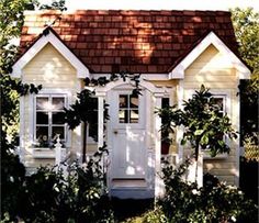 a small white house sitting in the middle of some bushes and trees with a brown shingled roof