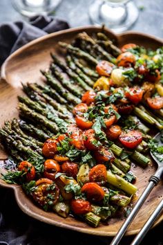 asparagus, tomatoes and other vegetables on a wooden plate with silverware next to wine glasses