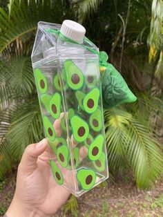 a hand holding up a plastic bottle with green and brown designs on the side, in front of some palm trees