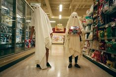 two people dressed in ghost costumes walking down a store aisle with shelves full of food