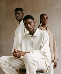 three men sitting on stools in front of a beige wall, one wearing a white shirt and pants