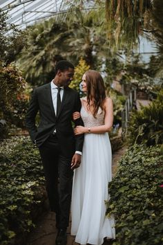 a man and woman in formal wear walking through the greenhouses with plants on either side