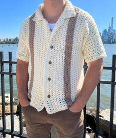 a man standing in front of a fence with his hands in his pockets, wearing a crocheted shirt and shorts