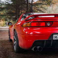 the rear end of a red sports car parked in a parking lot next to trees