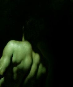 a man with no shirt on standing in the dark holding his hand up to his chest