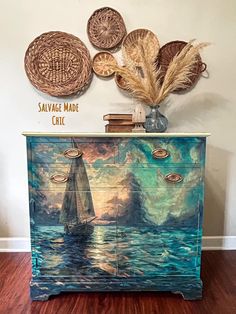 a painted dresser with decorative artwork on the top and bottom drawers, along with wicker wall hangings