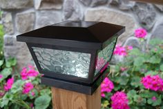 a close up of a light on a post near some plants and flowers in front of a brick wall